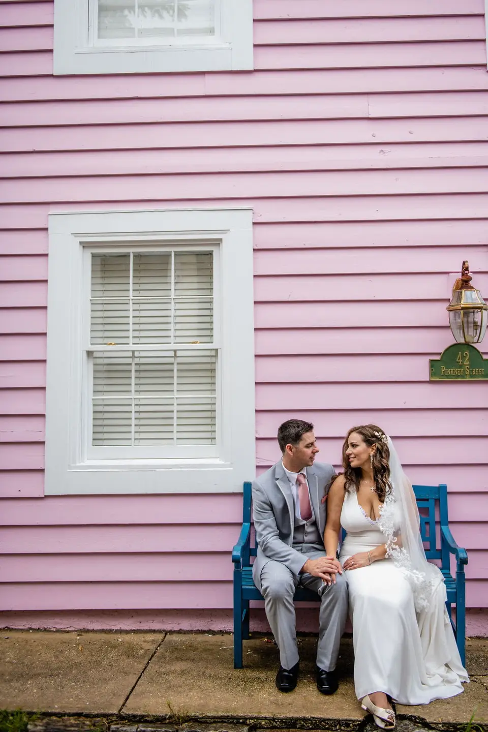 Bride and groom sitting on a blue bench in front of a pink clapboard building