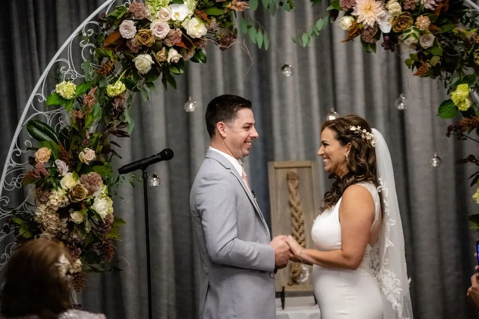 Bride and groom holding hands and smiling at each other during the ceremony under a floral arch