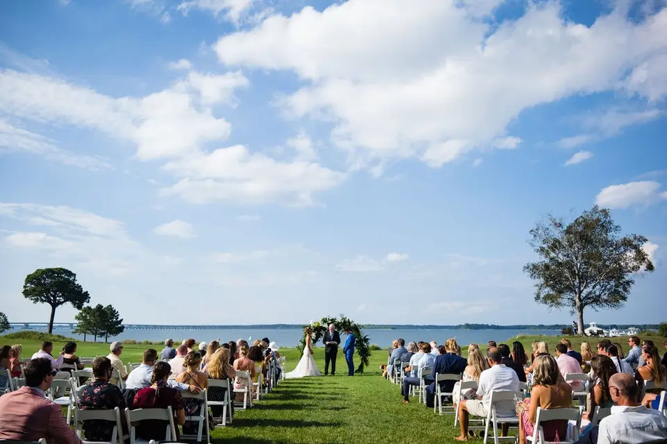 Outdoor wedding ceremony on a grassy lawn by the water with guests seated in white chairs