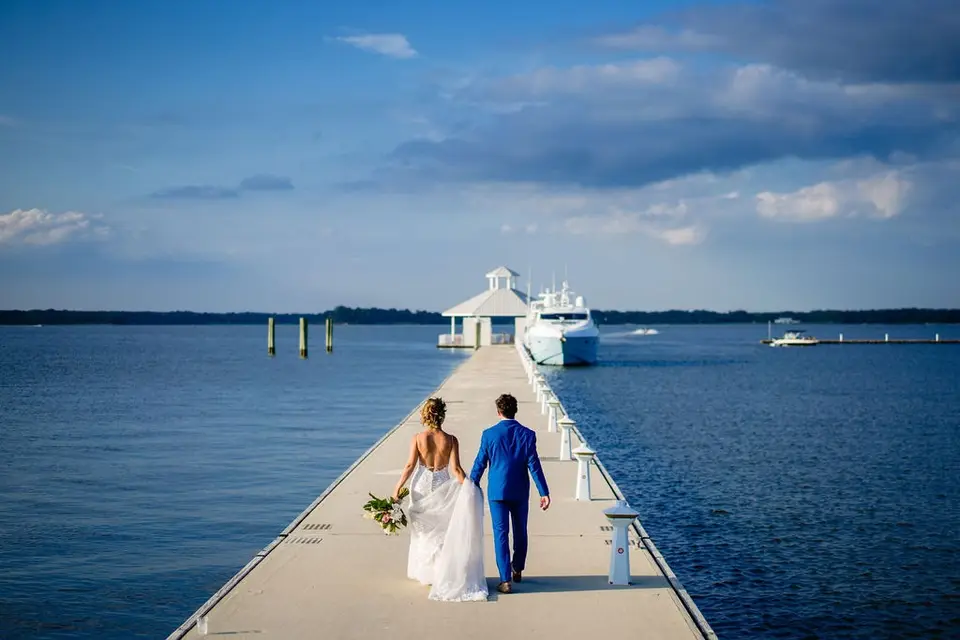 Bride and groom in a blue suit walking down a floating dock toward a gazebo and yacht