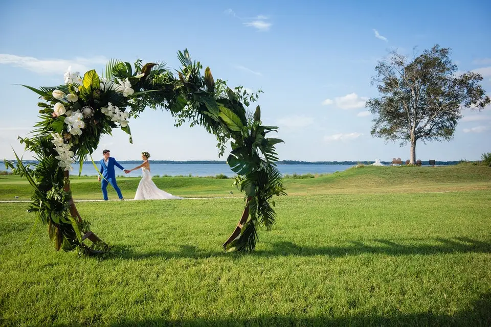 Couple holding hands under a round floral arch on a lawn overlooking the water