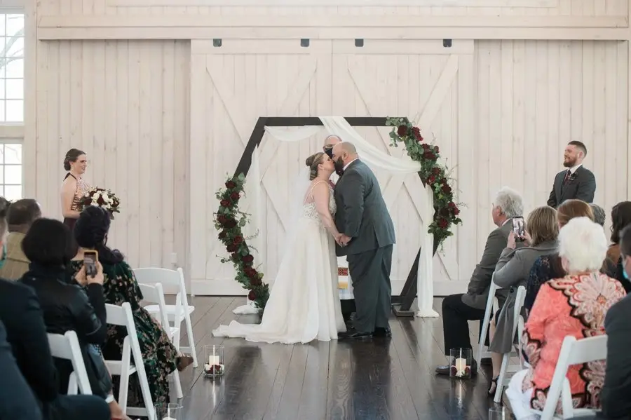 Bride and groom kissing under a hexagonal arch with red floral accents in a white barn venue as guests watch