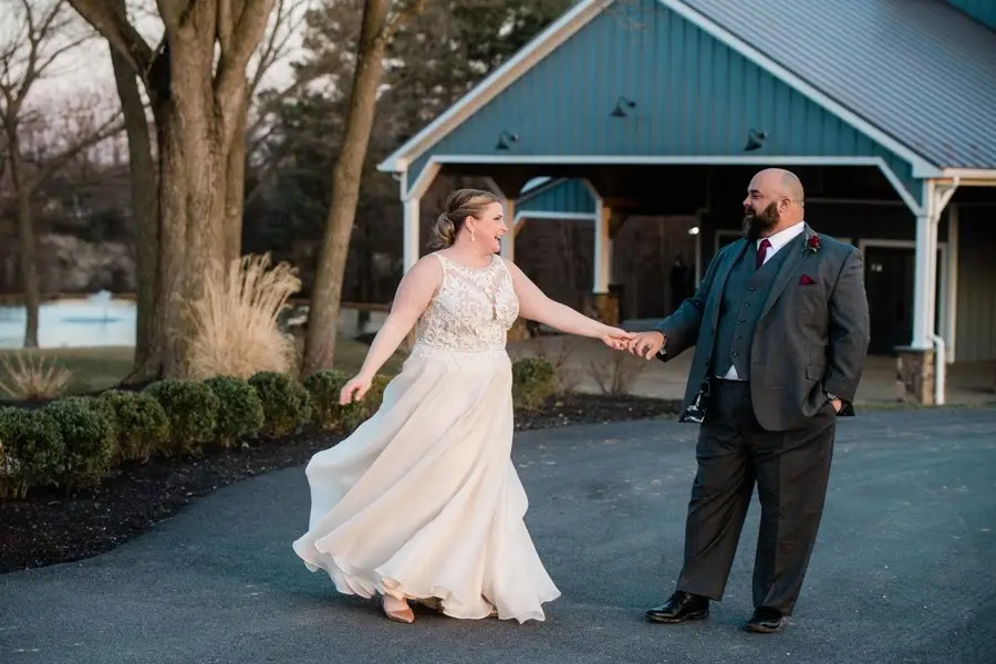 Bride and groom holding hands and smiling on a paved drive in front of a blue barn building