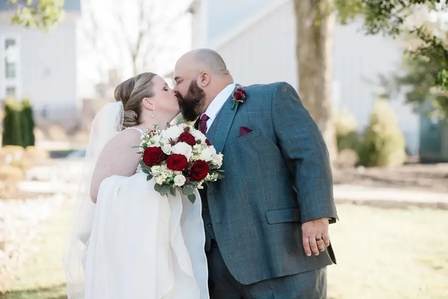Bride and groom kissing outdoors, holding a bouquet of red and white roses