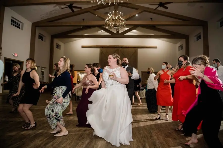 Bride in a white gown dancing with guests at a barn reception under a chandelier