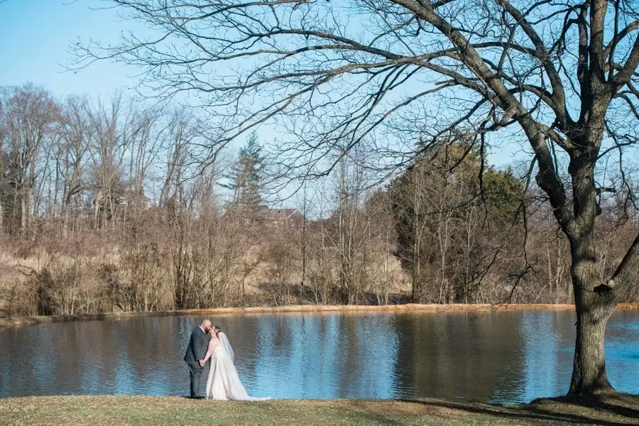 Bride and groom embracing on the shore of a small lake surrounded by bare trees