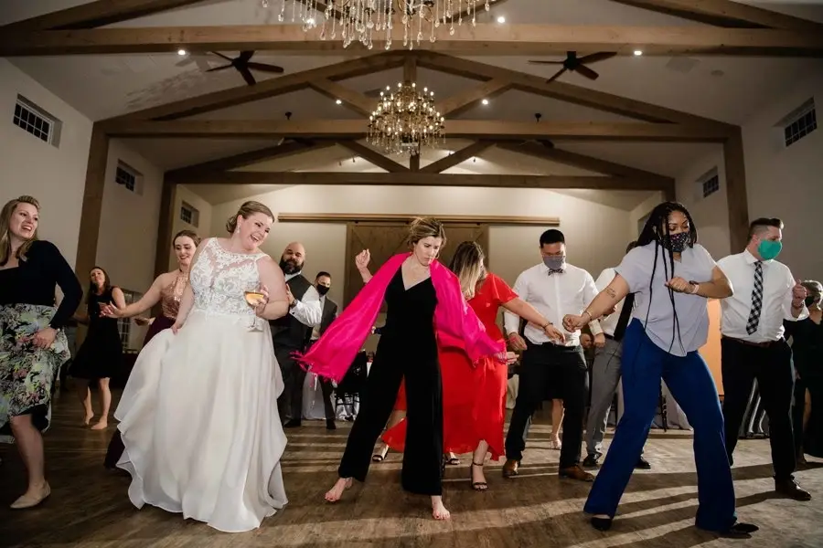 Wedding guests and the bride dancing together under a chandelier in a barn reception hall