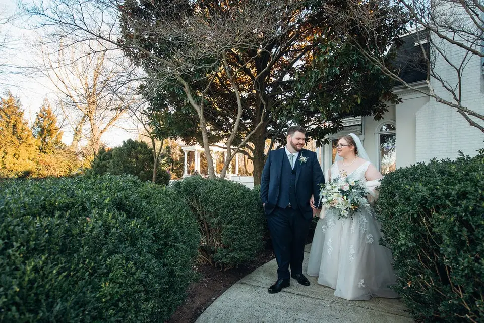 Groom and bride in a veil walking along a stone path between hedges, smiling at each other
