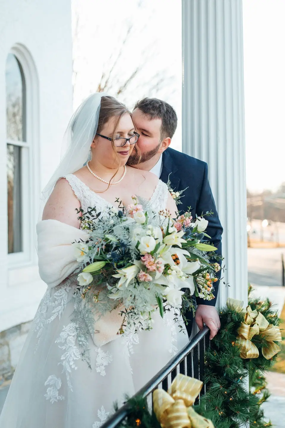 Bride in glasses and veil holding a large bouquet as the groom leans in behind her on a porch with a pine garland