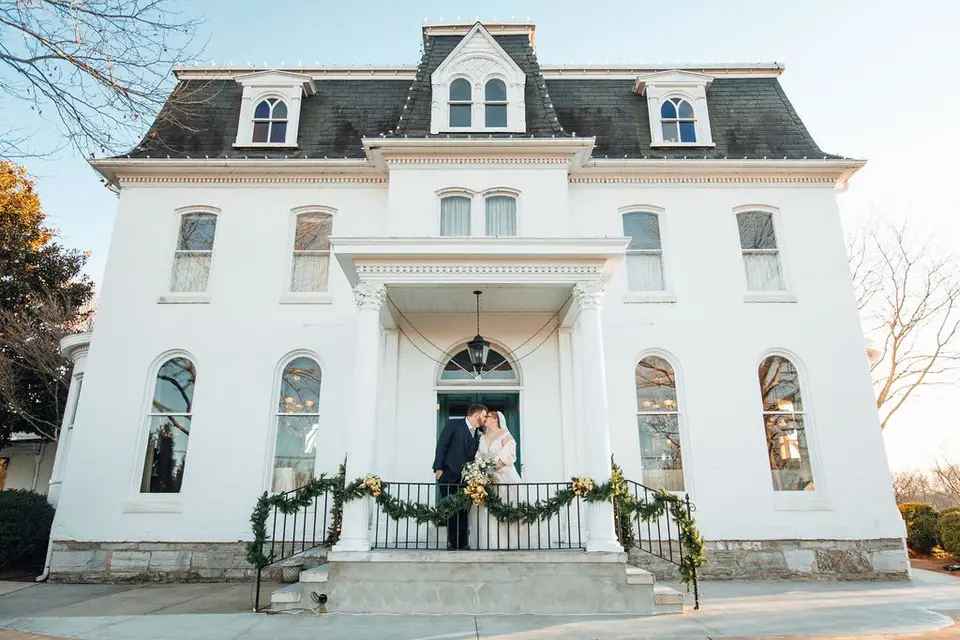 Bride and groom embracing in the doorway of a white mansion with a garland on the porch railing