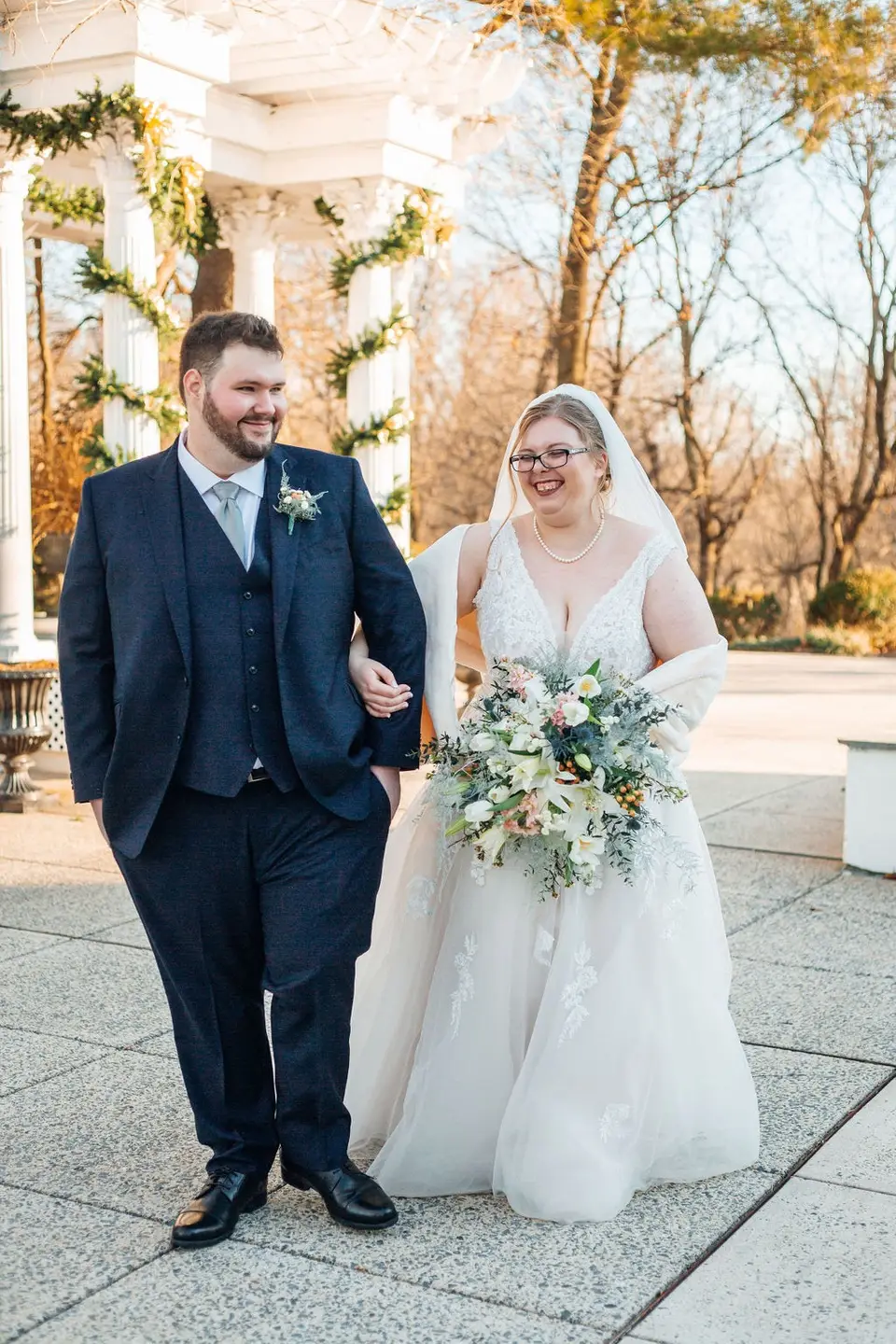 Smiling groom and bride in glasses with a bouquet walking arm in arm on a paved terrace