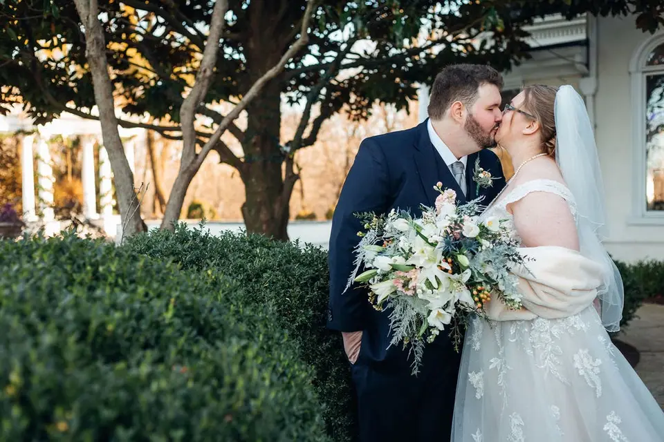 Groom kissing bride beside a bouquet outside near a tree and hedges, she wears a veil and wrap