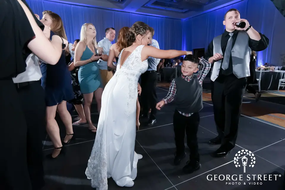 Bride dancing with a young boy on the dance floor as a man sings into a microphone in blue light