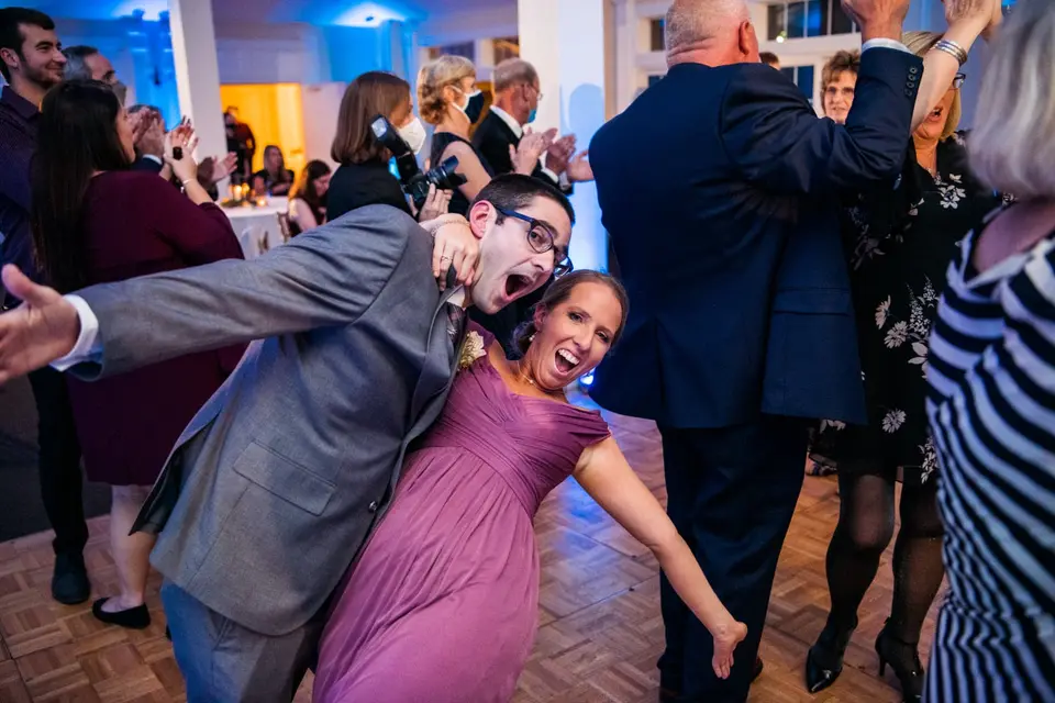 Man in glasses and a woman in a purple dress dance with arms outstretched at a reception