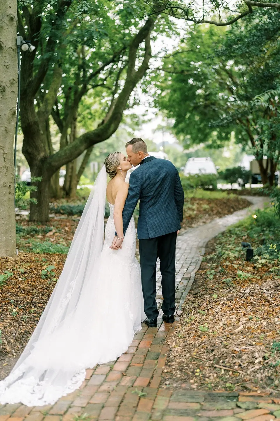 Bride and groom kiss on a brick path under shady trees, her long veil trailing behind