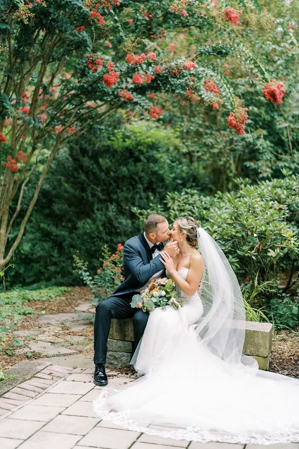 Groom and bride in a long veil kissing on a stone bench among shrubs with red blossoms