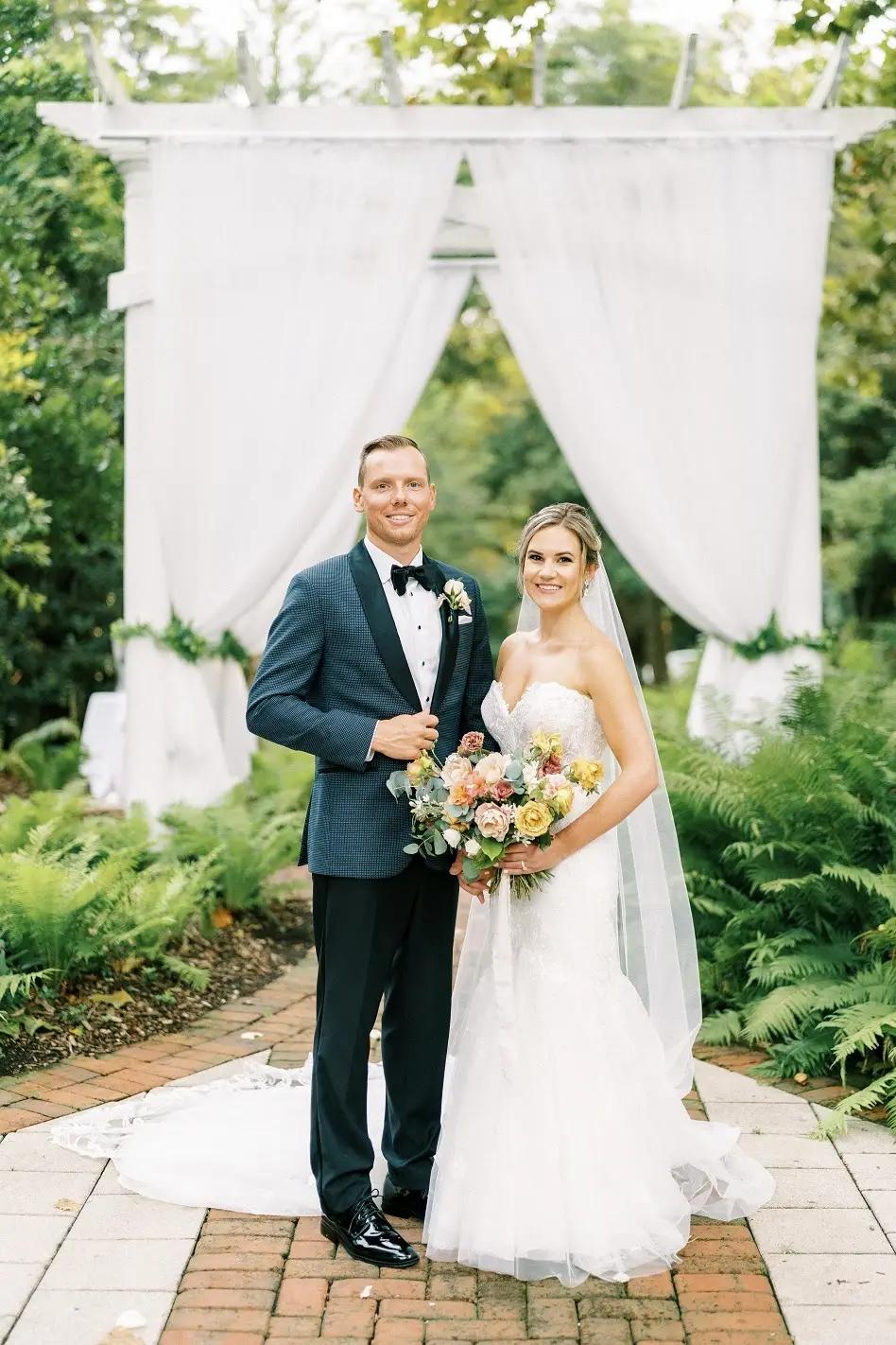 Bride holding a bouquet and groom in a teal jacket stand in front of a white draped ceremony arbor