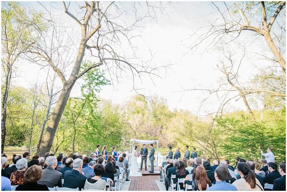Outdoor wedding ceremony at a floral arch with guests seated on both sides among spring trees