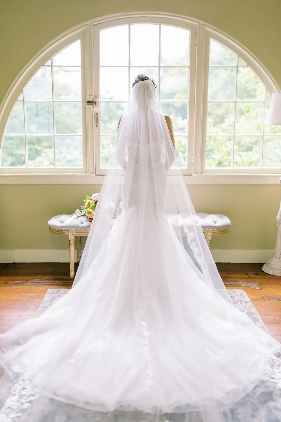 Bride in a long gown and cathedral veil facing an arched window with a small bouquet on a bench beside her