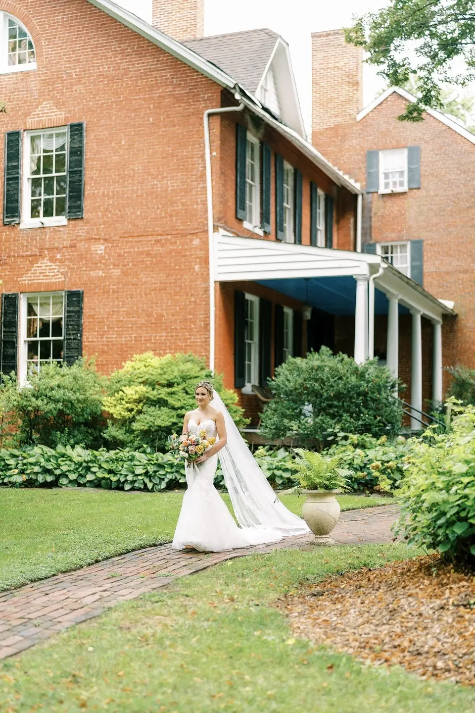 Bride in a long veil walking along a brick path toward a red brick house with green shutters