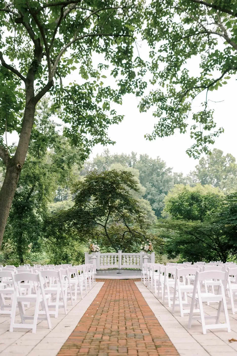 Rows of white folding chairs along a brick aisle leading to a garden ceremony spot