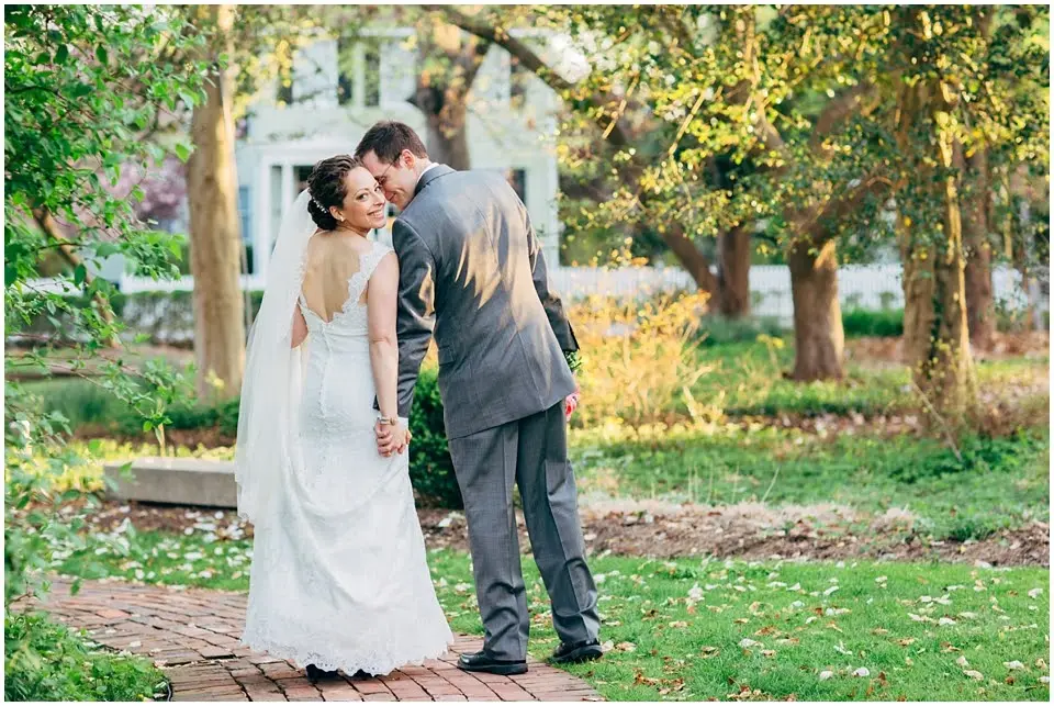 Groom in a gray suit and bride in a lace gown looking back over their shoulders as they walk through a garden