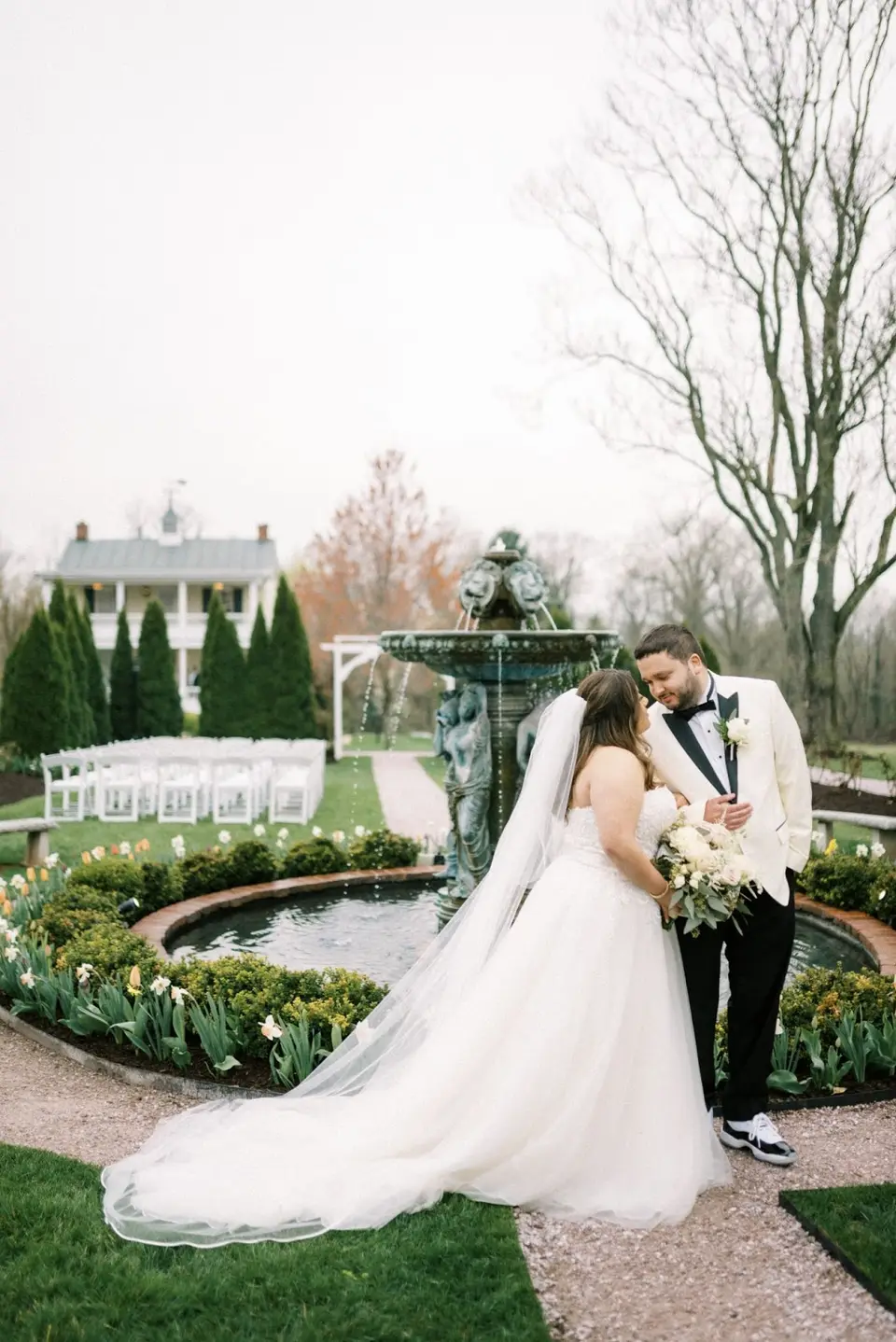 Bride and groom embracing beside a stone fountain in a landscaped garden, with white chairs set up behind