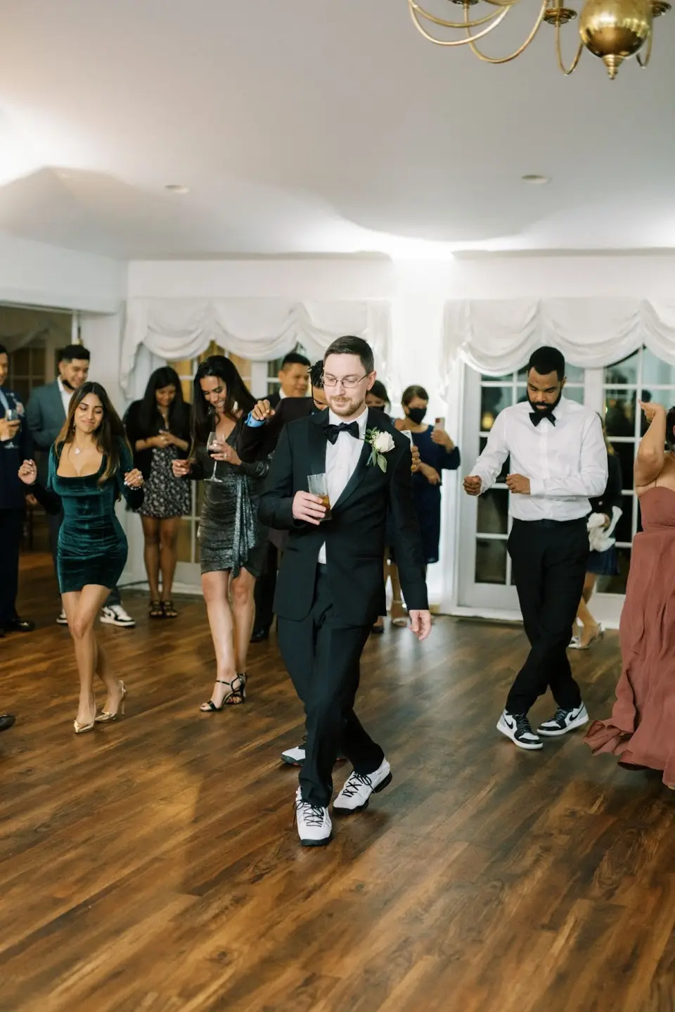 Wedding guests dancing on a wood floor at the reception, one man in a bow tie and white sneakers