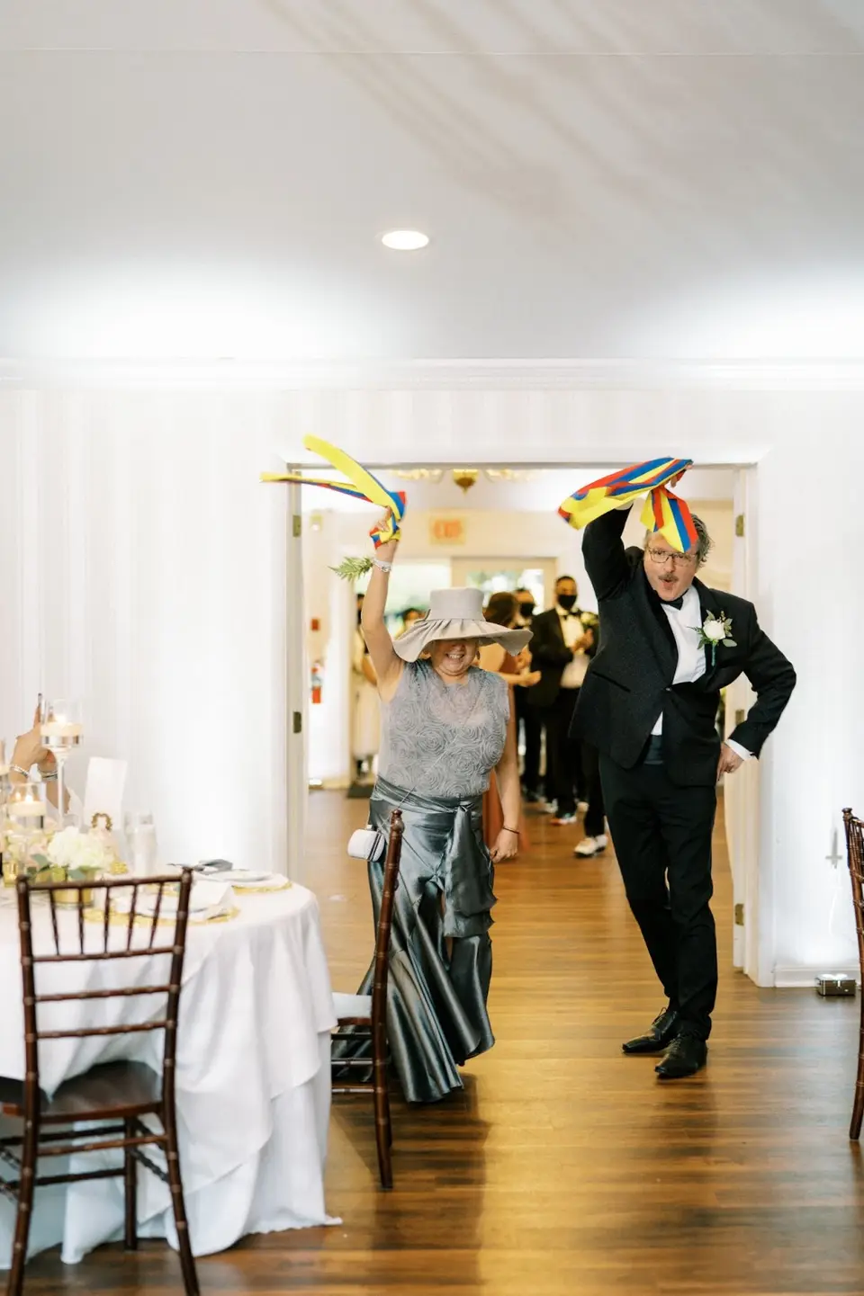 Older woman in a hat and man in a black suit waving colorful scarves as they enter the reception