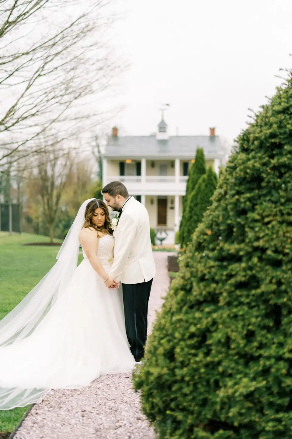Bride and groom embracing on a path with a white historic house and trimmed shrubs behind them