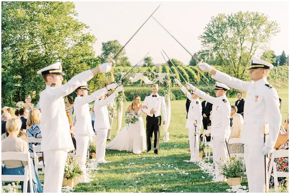 Bride and groom walking through an arch of raised sabres held by officers in white uniforms at a vineyard