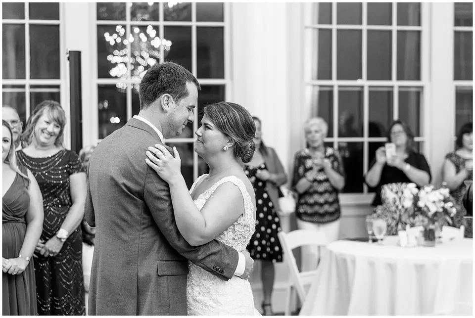 Bride and groom dancing close during their first dance as guests smile, black and white