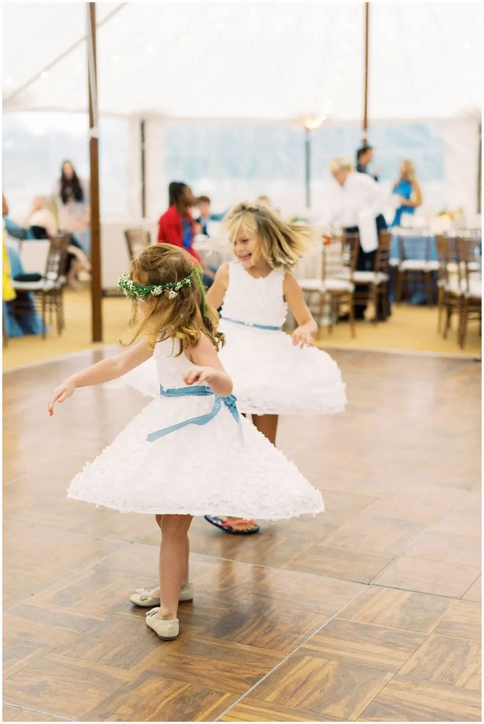 Two little girls in white dresses twirling on a wooden dance floor under a tent