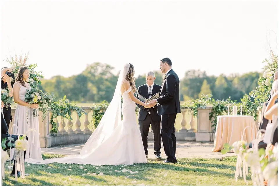 Bride and groom holding hands before an officiant at an outdoor ceremony with a stone balustrade