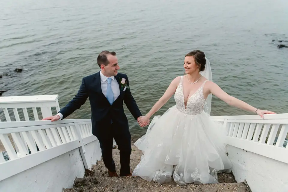 Groom holding the bride's hand as she stands on white steps leading down to the water at the Bay wedding