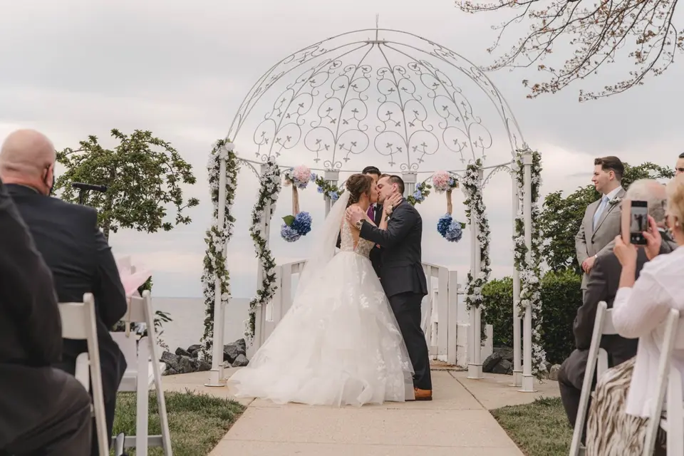 Bride and groom kissing at the altar under a white wrought-iron gazebo with blue hydrangeas as guests watch