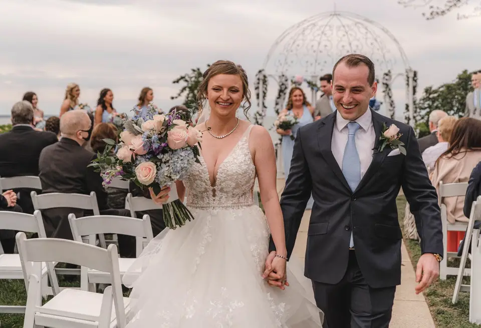 Allayne and Stephen holding hands and smiling as they walk up the ceremony aisle past seated guests