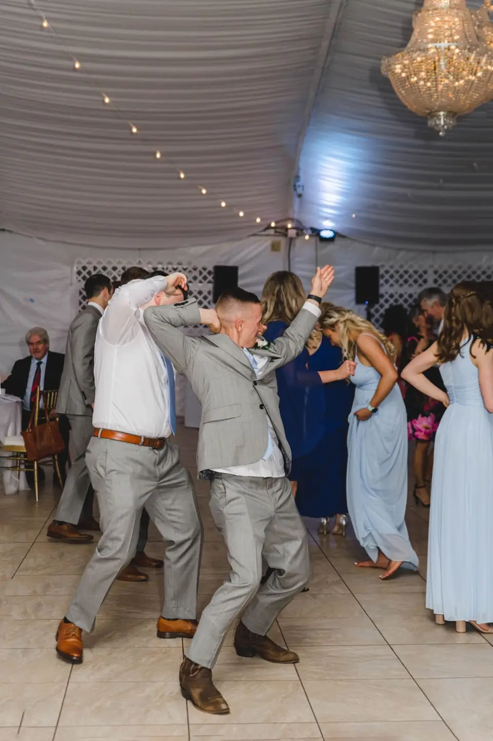Two men in gray suits dancing with arms raised on the dance floor under a white reception tent