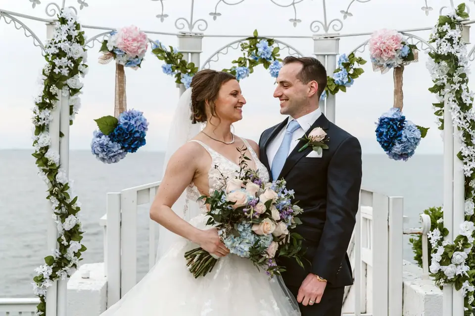 Allayne and Stephen smiling at each other under a floral-decorated white gazebo with the bay behind them