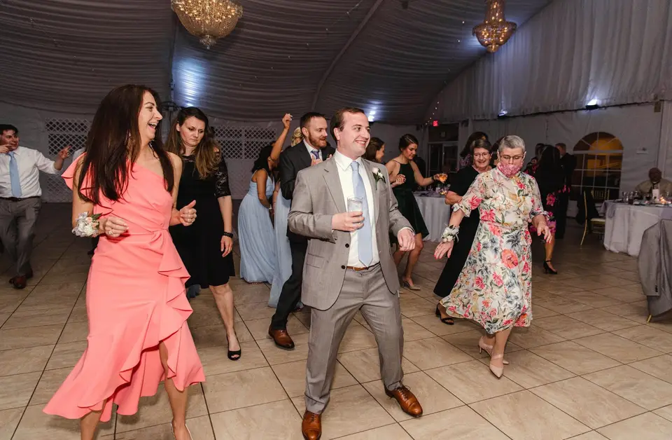 Guests dancing in a tent reception, including a man in a gray suit and women in pink and floral dresses