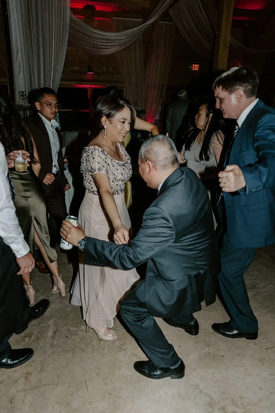 Man in a gray suit dancing low to the floor with a woman in a pink dress as other guests dance around them
