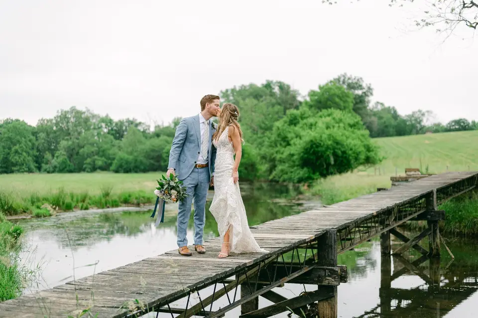 Allie and Pete kissing on an old wooden bridge over a pond, surrounded by green fields and trees
