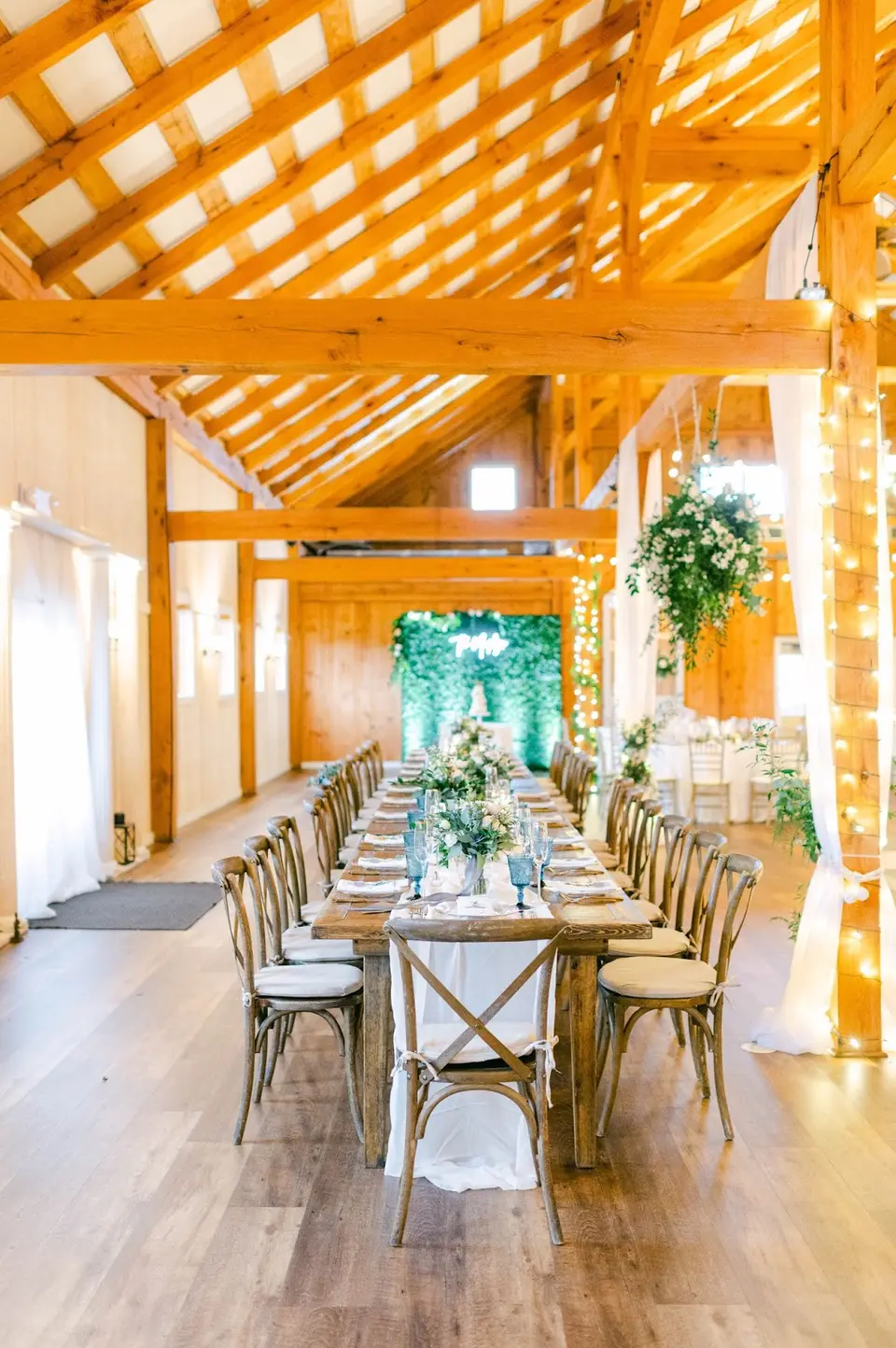 Long wooden farm table set with white flowers and greenery inside a rustic barn with exposed beams
