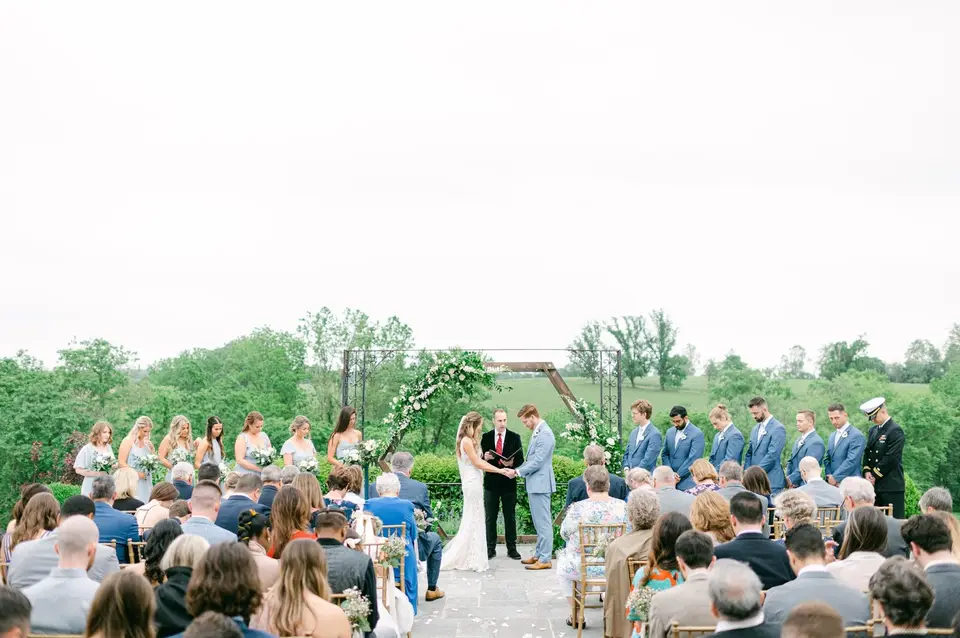 Allie and Pete exchanging vows outdoors with groomsmen, bridesmaids and seated guests in a green field
