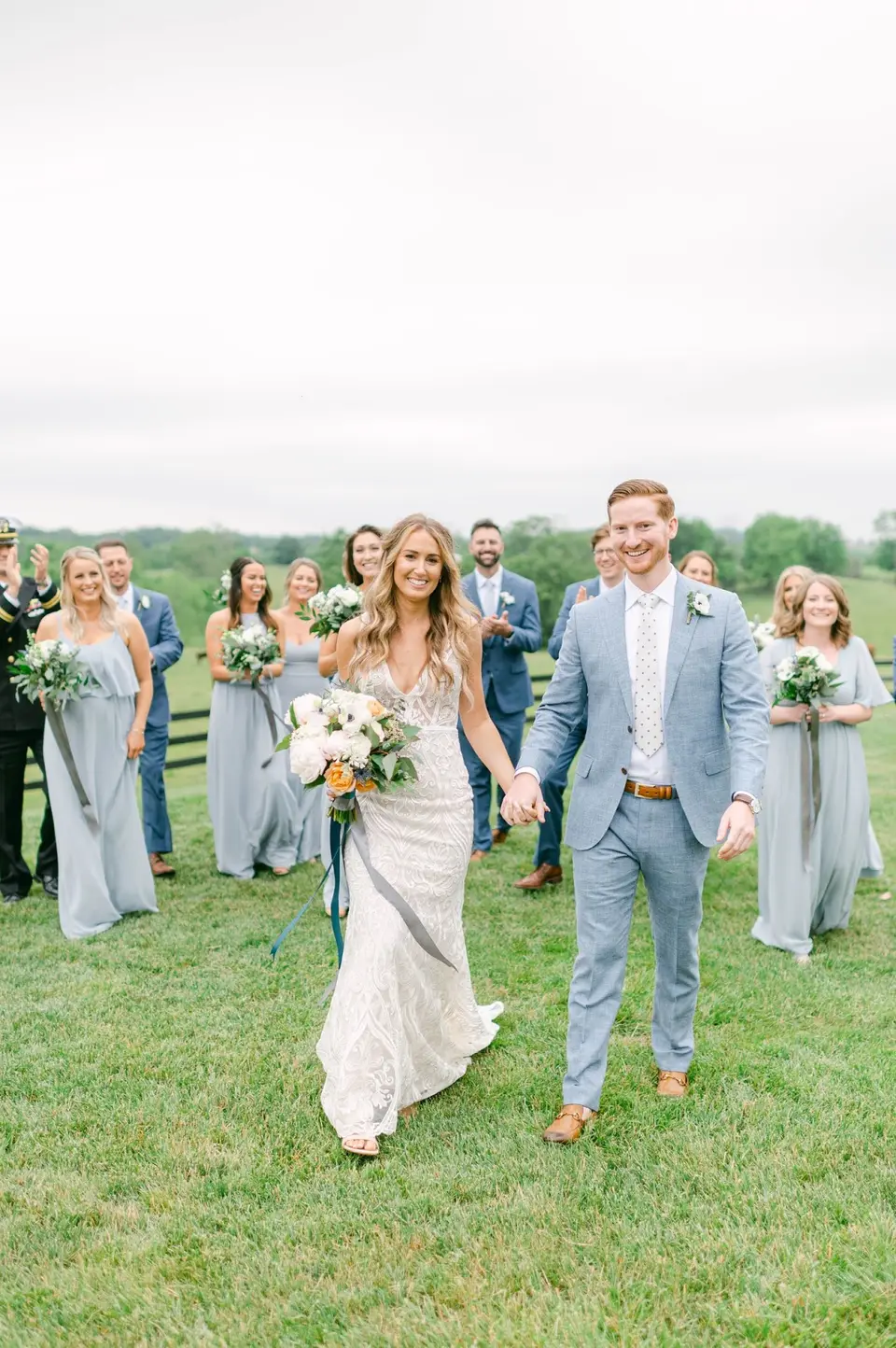 Bride and groom holding hands and smiling while the wedding party cheers behind them on the grass