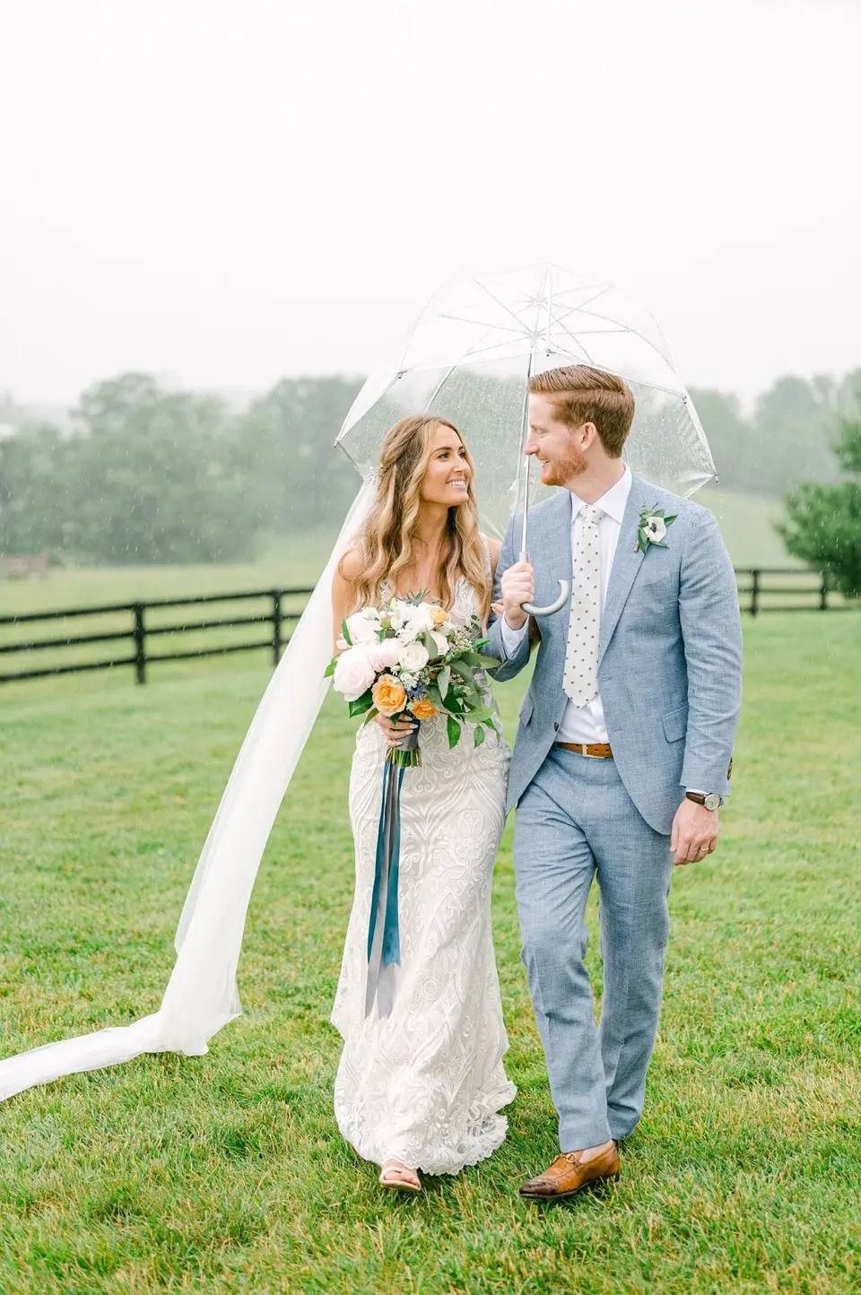 Bride in a lace gown and groom in a blue suit walking under a clear umbrella on a rainy lawn