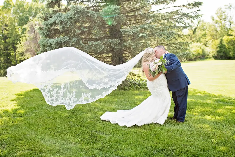 Bride and groom kiss on a lawn as her long lace veil blows out in the wind