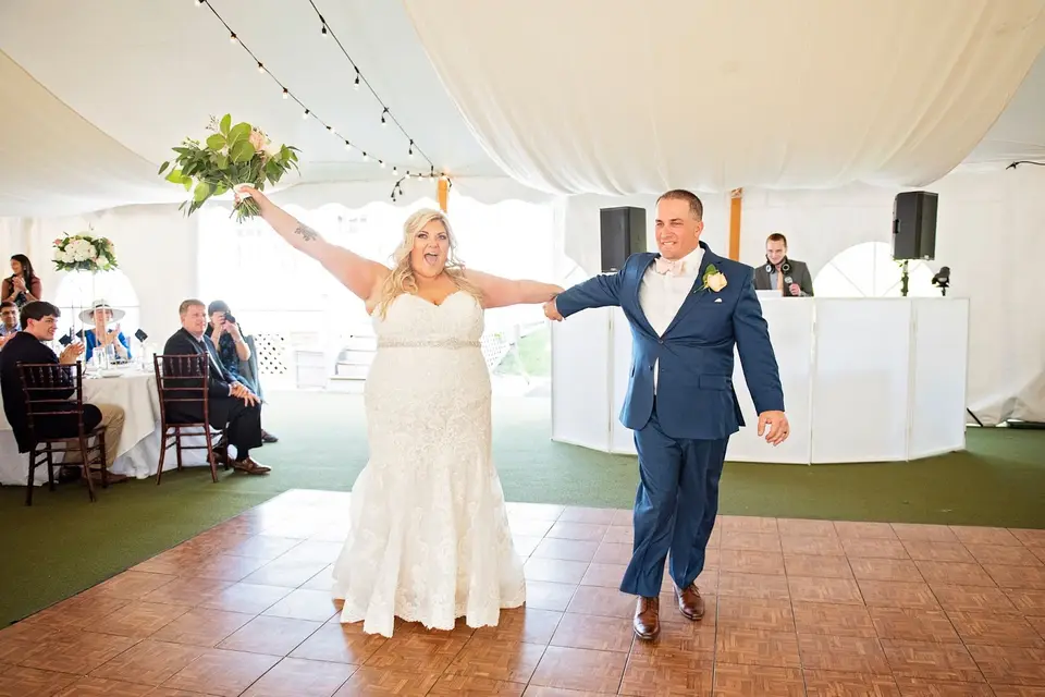 Bride raises her bouquet and groom gestures as they enter a tent reception with the DJ booth behind