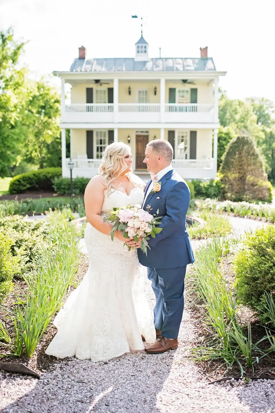 Bride and groom holding a pink bouquet on a garden path in front of a white historic house