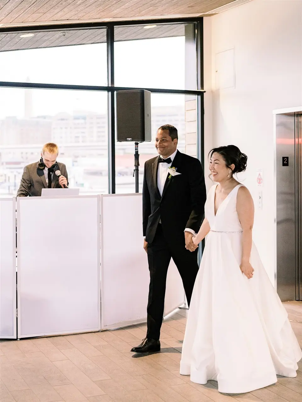 Bride and groom walk in holding hands as the DJ at a white-fronted booth speaks into a microphone
